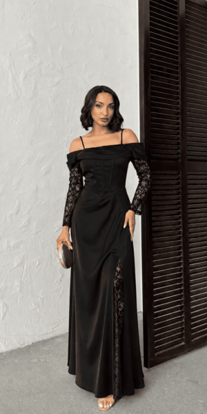 Robe de soirée longue noire en satin avec bustier corset, manches longues en dentelle et fente latérale, portée par un modèle avec une pochette dorée, vue de face en pied.